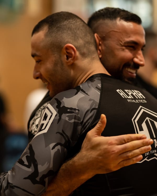 Zwei Mitglieder in Alpha-Rashguards umarmen sich lachend nach dem Training